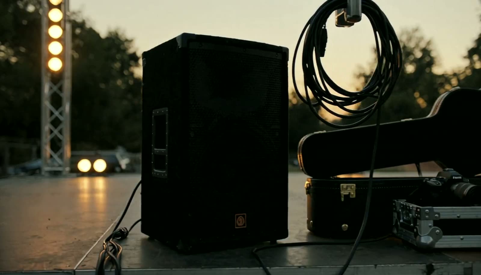 Evening venue preparation: a PA speaker and coiled cable on a dim stage, a guitar case open, a camera body resting on a road case.